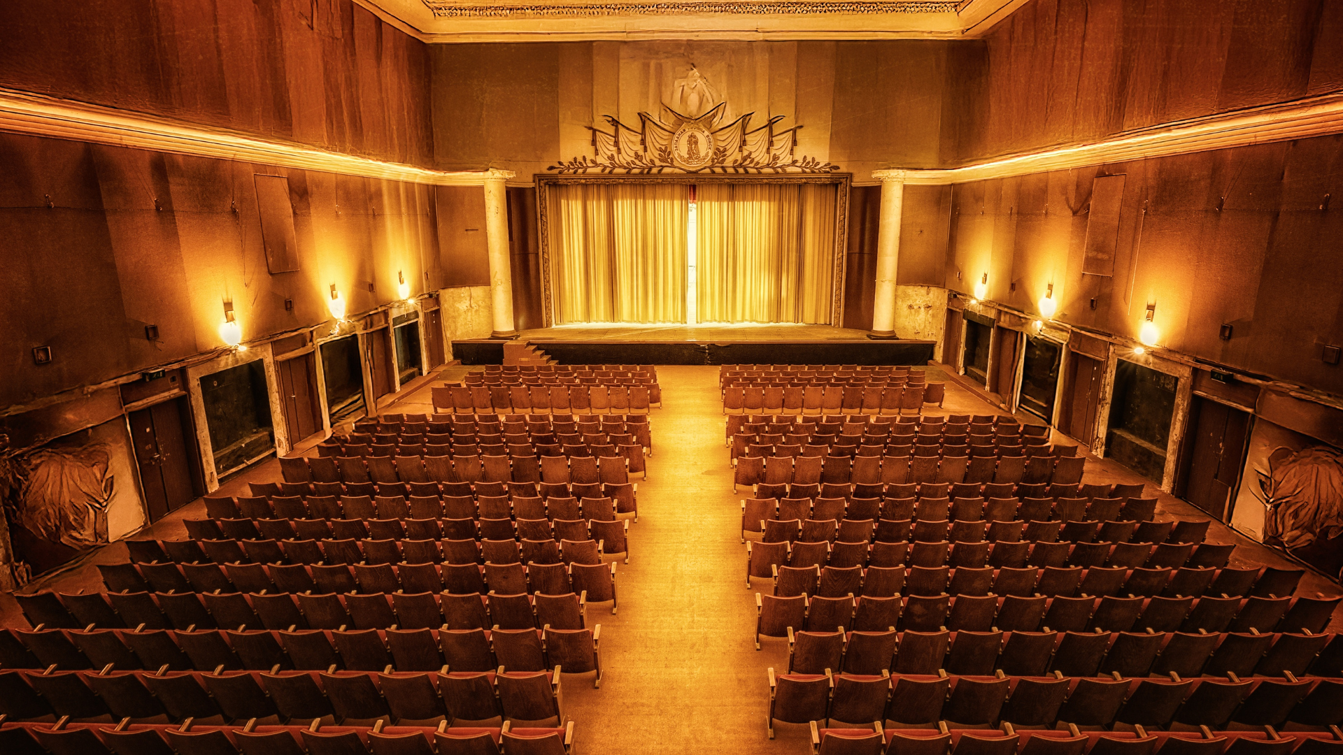 A single ghost light glowing in the center of a dark, empty stage at dusk, a chair turned to face it from the house.