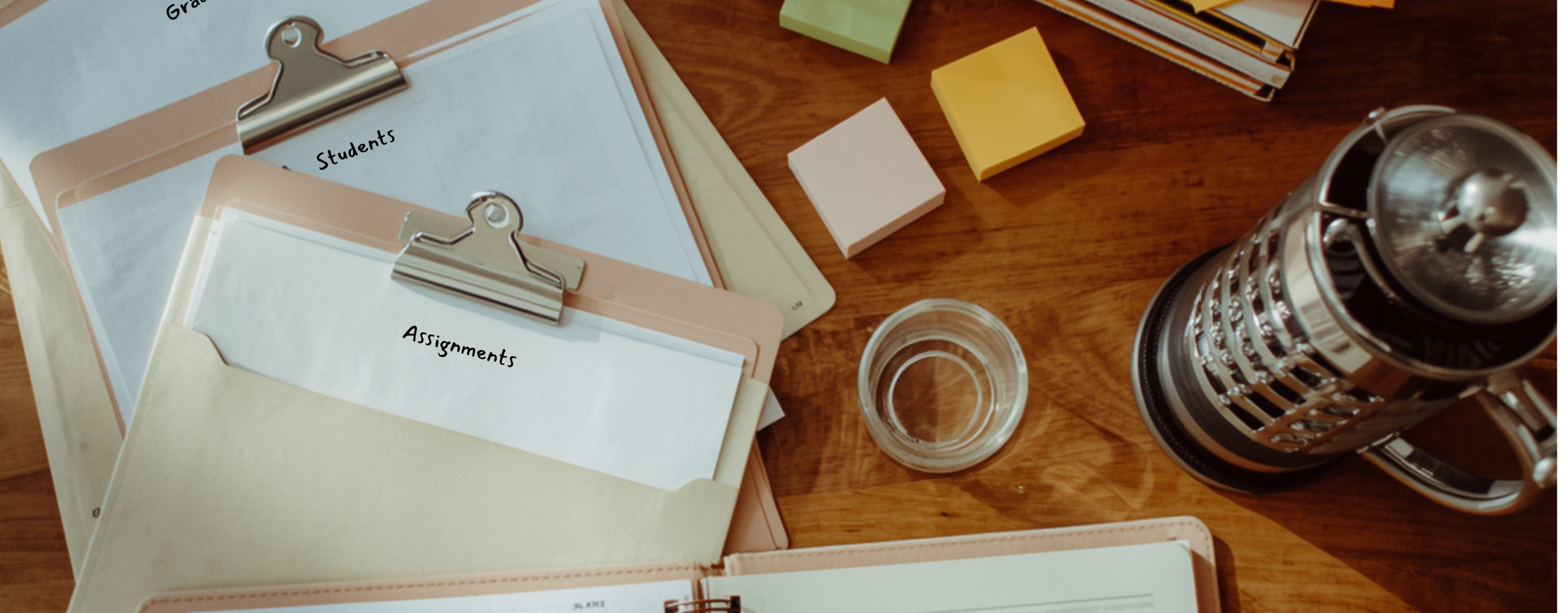 A head tutor&rsquo;s prep table overhead. Assignment folders fanned out, a stack of practice tests, color-coded sticky notes, a French press, an open notebook with a session-plan sketched in pencil.